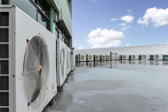 Rows of white air conditioning units on a wet rooftop under a blue sky with clouds.