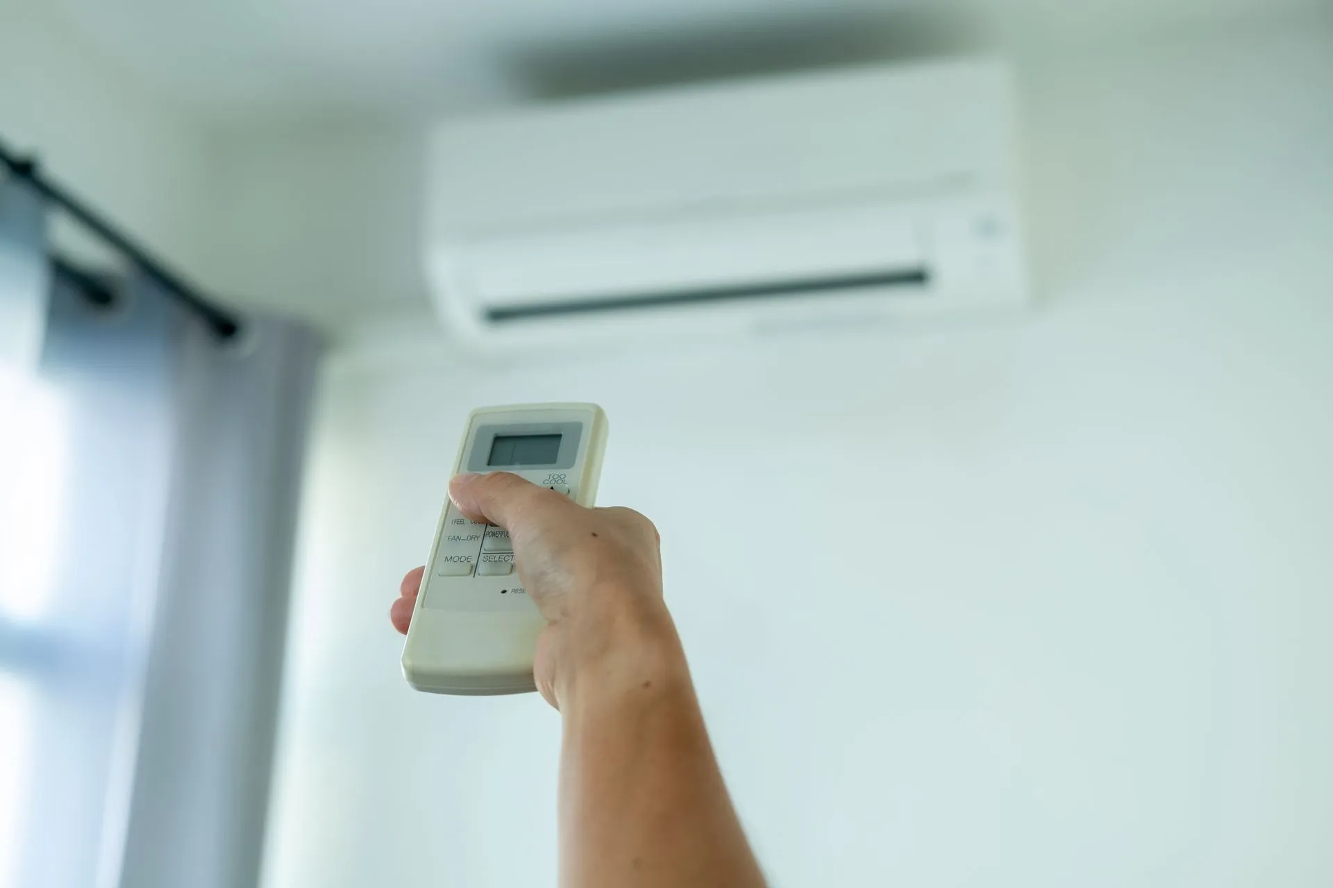 Hand holding AC remote, aiming towards a wall-mounted air conditioner.
