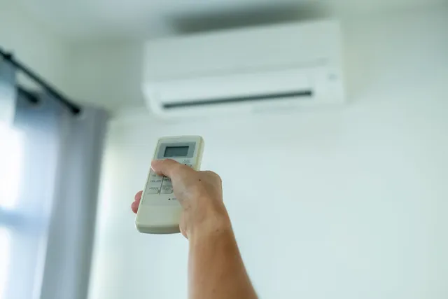 Hand holding AC remote, aiming towards a wall-mounted air conditioner.