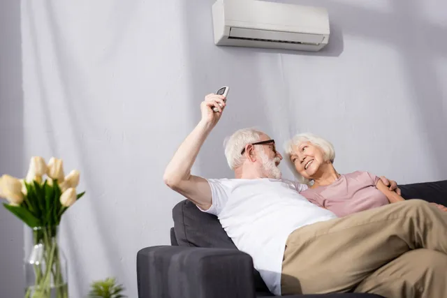 An elderly couple is sitting on a couch under an air conditioner.
